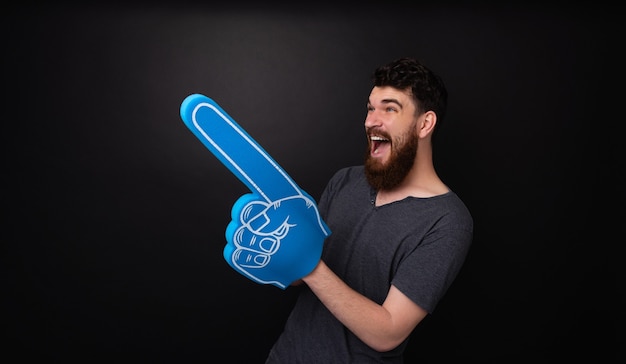 Photo d'un homme barbu avec un grand gant de ventilateur bleu pointant vers l'extérieur debout sur fond sombre