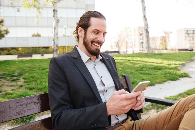 Photo d'un homme barbu joyeux de 30 ans en costume d'affaires tenant et utilisant un téléphone mobile, assis sur un banc dans le parc de la ville pendant la journée ensoleillée