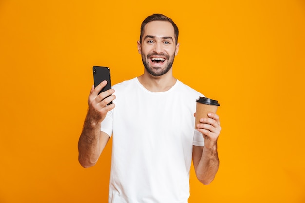 Photo d'un homme brune de 30 ans en tenue décontractée tenant un téléphone portable et un café à emporter, isolé