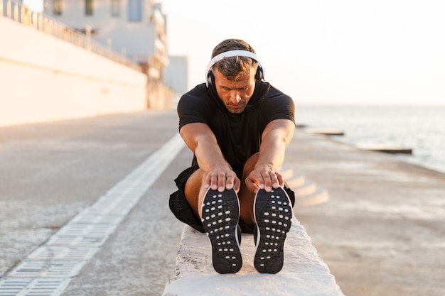 Photo d'un homme caucasien en bonne santé de 30 ans en survêtement s'échauffant et s'étendant les jambes au large de la côte, tout en écoutant de la musique via des écouteurs sans fil pendant le lever du soleil