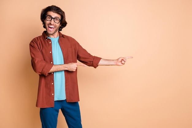 Photo d'un homme fou drôle indiquer un espace vide du doigt porter des lunettes t-shirt marron isolé sur fond de couleur beige