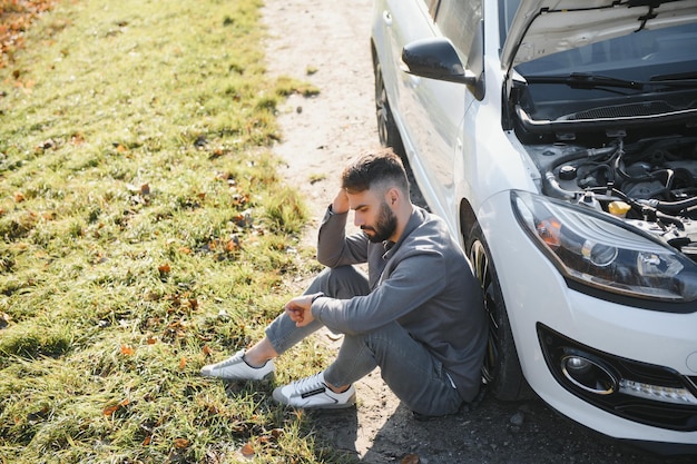 Photo d'un homme frustré assis à côté d'une voiture cassée avec le capot ouvert