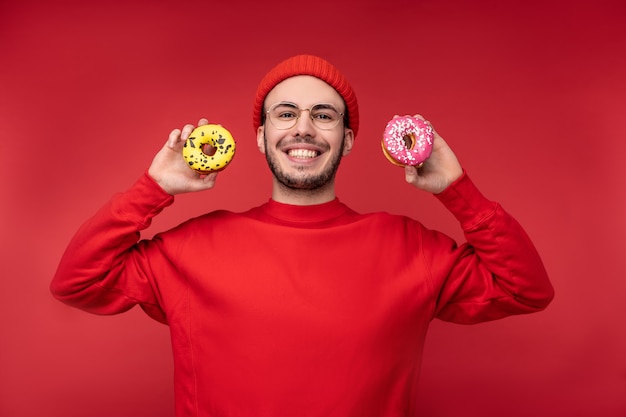 Photo d'un homme heureux avec un beau sourire et une barbe dans des lunettes et des vêtements rouges. Profitez de manger des beignets de bonbons, isolés sur fond rouge.