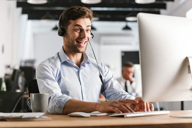 Photo de l'homme heureux opérateur 20s portant des vêtements de bureau et un casque, assis par ordinateur dans le centre d'appels