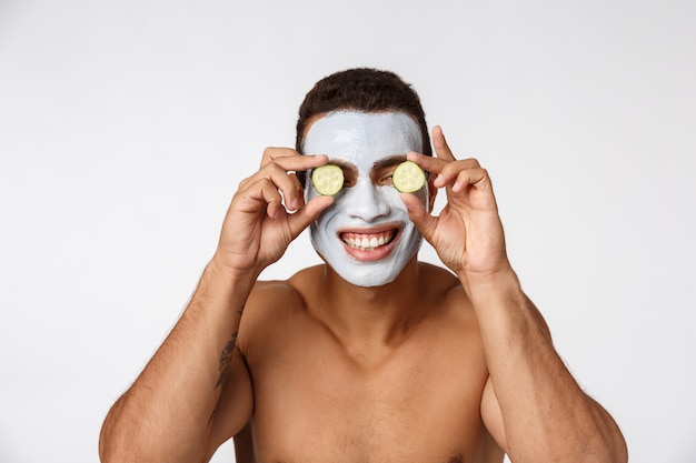 Photo d'un homme joyeux dans un masque cosmétique facial se moquant de concombre isolé sur blanc