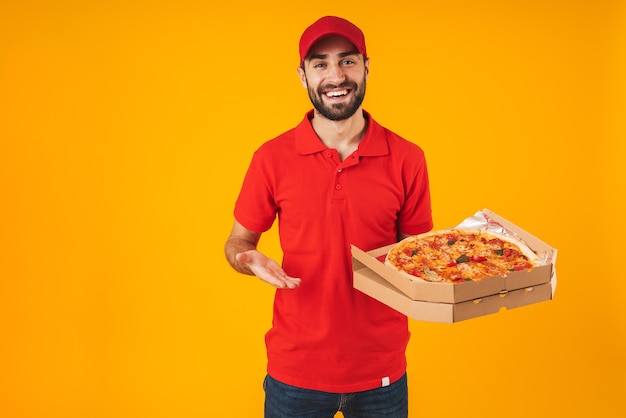 Photo d'un homme joyeux et heureux en uniforme rouge souriant et tenant une boîte à pizza isolée sur jaune
