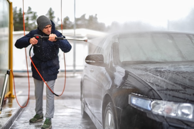 Une photo d'un homme lavant une voiture sous haute pression d'eau à l'extérieur pendant la saison froide.