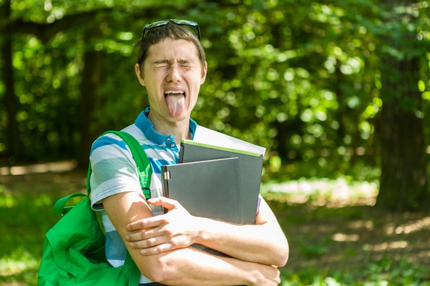 Photo d'un homme montrant la langue avec un ordinateur portable, des livres et un sac à dos dans un parc d'été