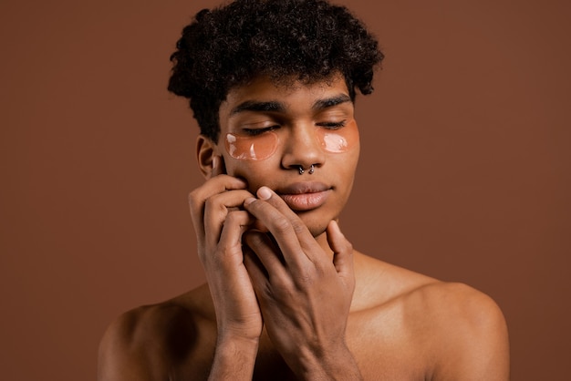 Photo d'un homme noir attrayant avec des piercings et des cache-yeux sur les yeux baissés et touche son visage. Torse nu, fond de couleur marron isolé.