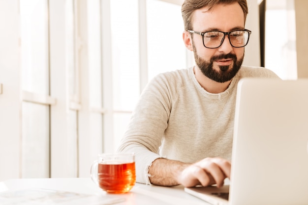 Photo d'un homme non rasé attrayant portant des lunettes, buvant du thé dans le verre, tout en travaillant ou en naviguant sur Internet sur un ordinateur portable au bureau