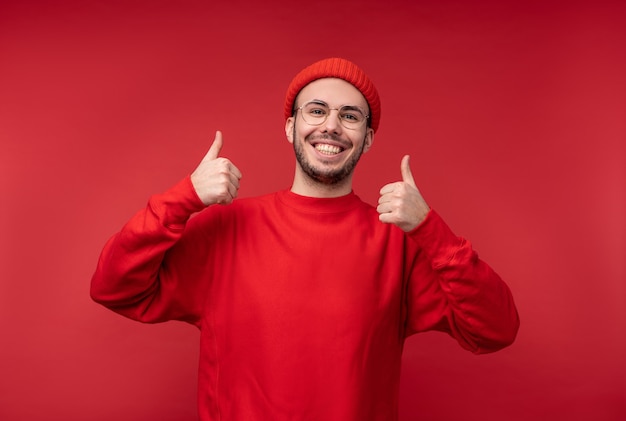 Photo d'un homme séduisant avec barbe à lunettes et vêtements rouges. Homme donne le pouce vers le haut et sourit, isolé sur fond rouge.