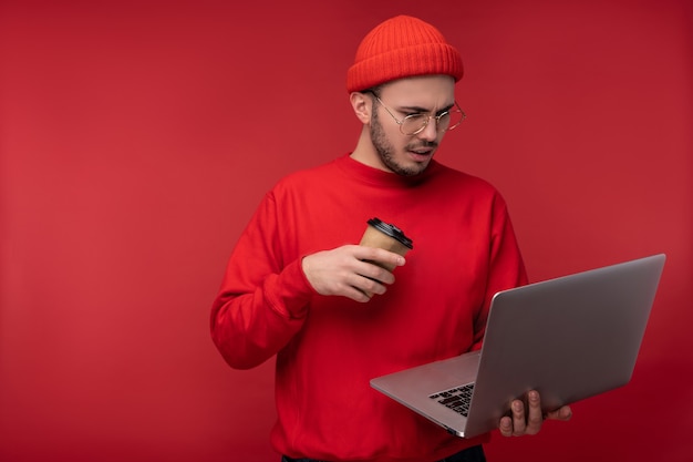 Photo d'un homme séduisant avec barbe à lunettes et vêtements rouges. Homme réfléchi tient un ordinateur portable et du café, isolé sur fond rouge.