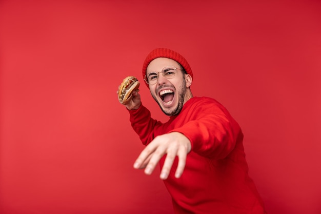 Photo d'un homme séduisant avec barbe à lunettes et vêtements rouges. Mâle détient un hamburger et va le lancer, isolé sur fond rouge.