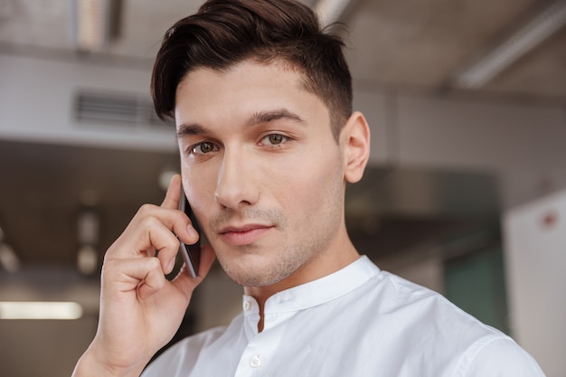 Photo d'un homme sérieux vêtu d'une chemise blanche parlant au téléphone à l'intérieur. Cotravail. En regardant la caméra.
