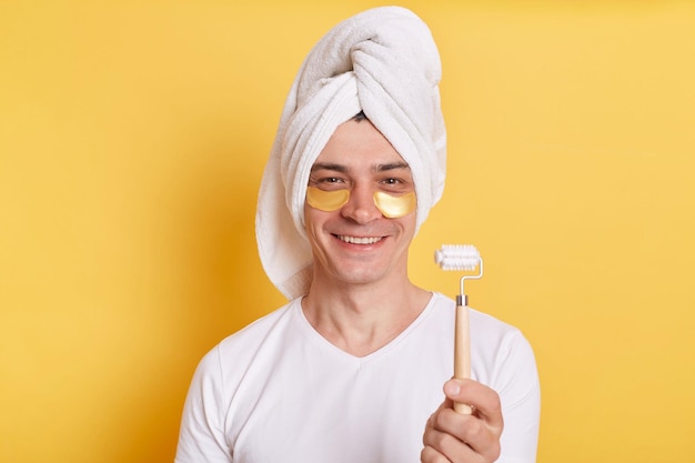 Photo d'un homme souriant et satisfait portant un t-shirt blanc et enveloppé dans une serviette tenant un rouleau de massage faisant des procédures de soins de la peau debout isolé sur fond jaune