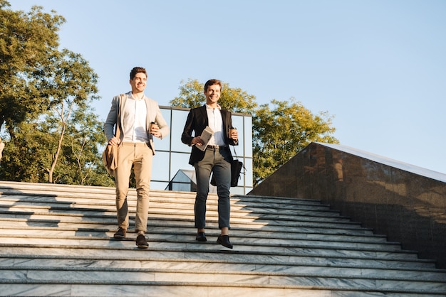 Photo d'hommes d'affaires européens en costume marchant en plein air avec du café à emporter, pendant la journée ensoleillée