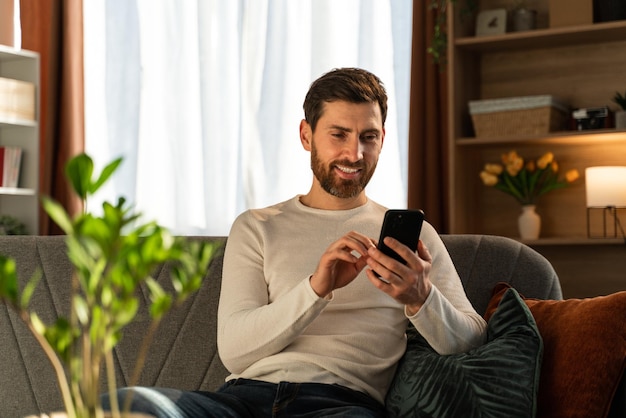 Photo horizontale du jeune homme d'affaires heureux faisant du travail indépendant dans sa chambre et tapant des messages sur le smartphone