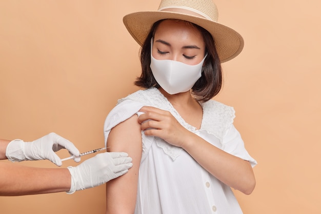 Une photo horizontale d'une femme asiatique portant un masque stérile de protection pour empêcher la propagation du coronavirus reçoit une injection de vaccin dans les poses d'épaule contre un mur beige