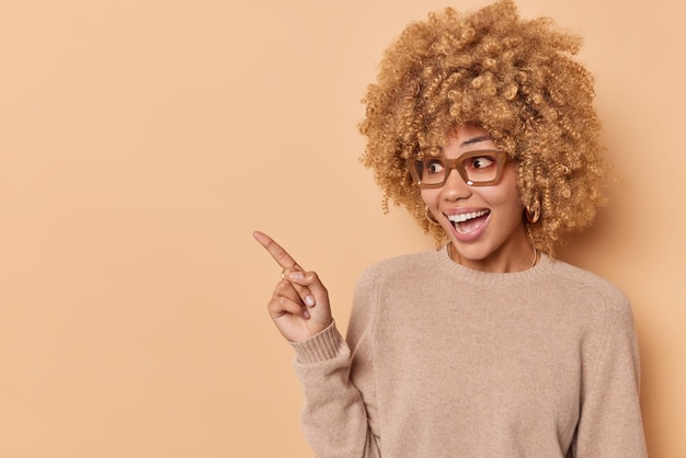 Une photo horizontale d'une femme aux cheveux bouclés gaie pointe vers l'extérieur sur un espace vide montre quelque chose de bien démontre une publicité ou une remise vêtue d'un pull décontracté isolé sur fond beige.