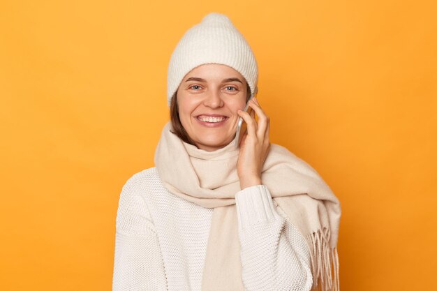 Photo horizontale d'une femme joyeuse et souriante heureuse portant un pull blanc, un chapeau et une écharpe posant isolé sur fond jaune debout regardant la caméra parler sur un téléphone mobile