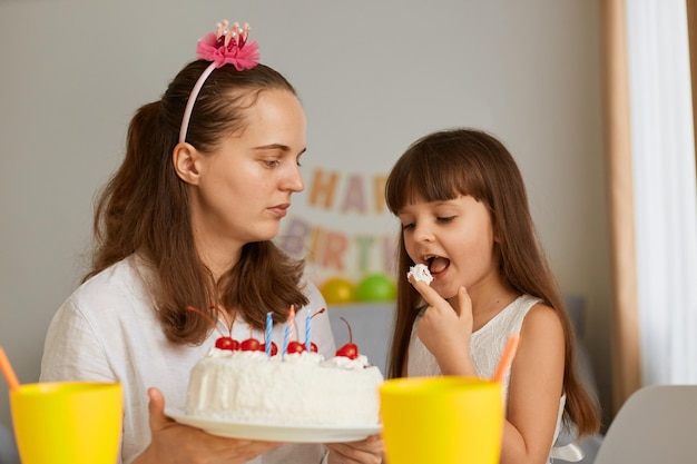 Photo horizontale d'une mère avec sa fille célébrant son anniversaire, petite fille léchant le doigt dégustant un délicieux dessert festif, fête à la maison, joyeuses fêtes en famille.