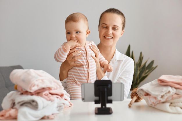 Photo d'intérieur d'une blogueuse souriante et satisfaite assise à table avec un enfant en bas âge et enregistrant une vidéo pour son vlog, faisant du contenu avec sa fille, posant devant un téléphone sur un trépied.