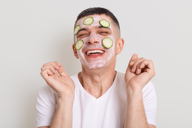 Photo d'intérieur d'un charmant homme gentil et charmant portant un t-shirt blanc faisant des procédures de cosmétologie à la maison debout avec les bras levés et riant posant isolé sur fond gris