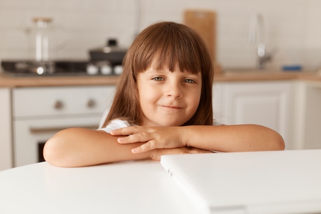 Photo d'intérieur d'une charmante petite fille d'âge préscolaire aux cheveux noirs assise à table avec un cahier plié, regardant la caméra, posant à la maison dans une pièce lumineuse avec un ensemble de cuisine en arrière-plan.