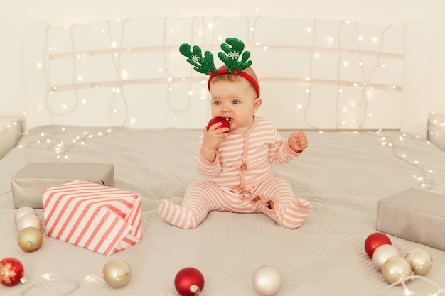 Photo d'intérieur d'une charmante petite fille portant un dormeur rayé assis sur un lit avec une décoration de Noël, mordant une boule rouge, explorant le monde autour, célébrant le nouvel an.