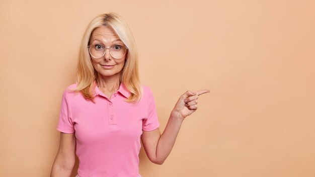 Une Photo D'intérieur D'une Femme Européenne Blonde Surprise Pointe Du Doigt Sur Un Espace Vide Présente Une Offre Spéciale Portant Des Lunettes Transparentes Et Un T-shirt Rose Isolé Sur Un Mur Beige