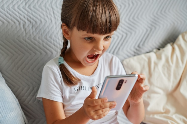 Photo d'intérieur d'une fille agressive portant un t-shirt décontracté blanc et ayant les cheveux noirs, tenant un téléphone intelligent dans les mains, étant en colère de perdre son niveau, criant de colère.