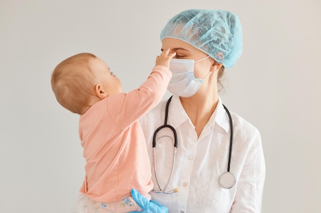 Photo d'intérieur d'une gentille jeune pédiatre adulte portant un uniforme médical, tenant une petite fille, examinant la santé de l'enfant à la clinique, une petite fille touchant une infirmière.