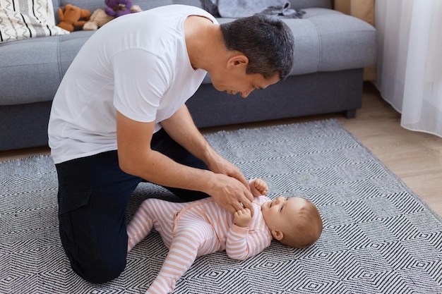 Photo d'intérieur d'un homme brune espiègle portant un t-shirt blanc jouant avec sa petite fille à la maison, un enfant mignon en dormeur allongé sur le sol et regardant son père.