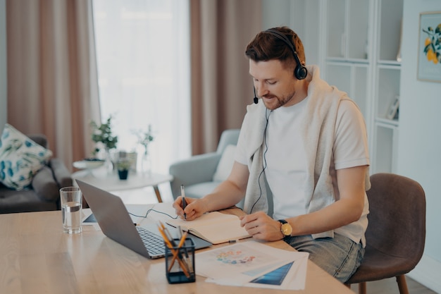 Une photo d'intérieur d'un homme heureux prend des notes tout en écoutant des cours en ligne étudie à distance depuis la maison est assise au bureau avec des papiers d'ordinateur portable modernes porte des écouteurs a une conférence Web ou une réunion virtuelle