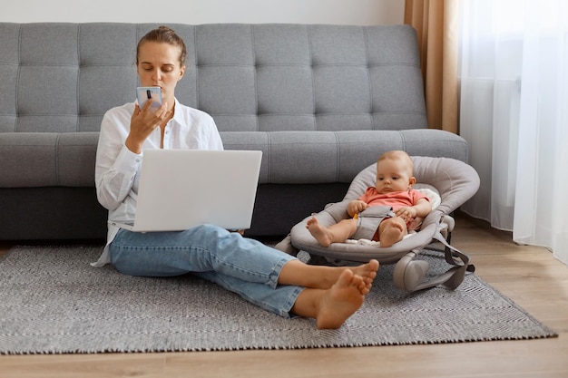 Photo d'intérieur d'une jeune femme adulte concentrée portant une chemise blanche travaillant sur ordinateur et passant du temps avec son bébé dans un videur, enregistrant un message vocal.