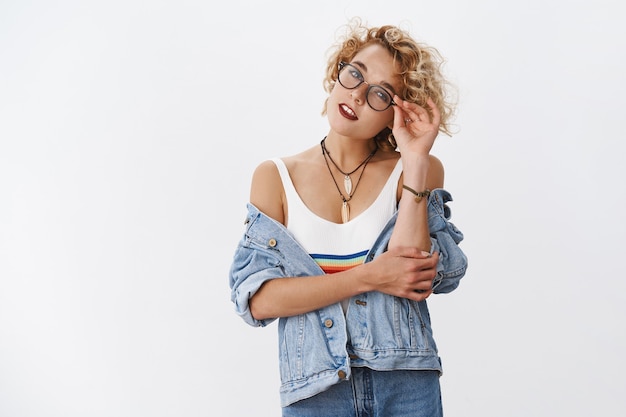 Photo d'intérieur d'une jeune femme élégante et sensuelle en veste en jean et haut tendance avec des lunettes de contrôle arc-en-ciel inclinant la tête et la bouche ouverte séduisant et flirtant sur un mur blanc