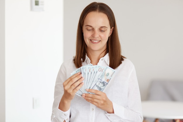 Photo d'intérieur d'une jolie femme aux cheveux noirs vêtue d'une chemise blanche de style décontracté, tenant un ventilateur d'argent dans les mains, comptant les billets de banque avec une expression faciale heureuse.