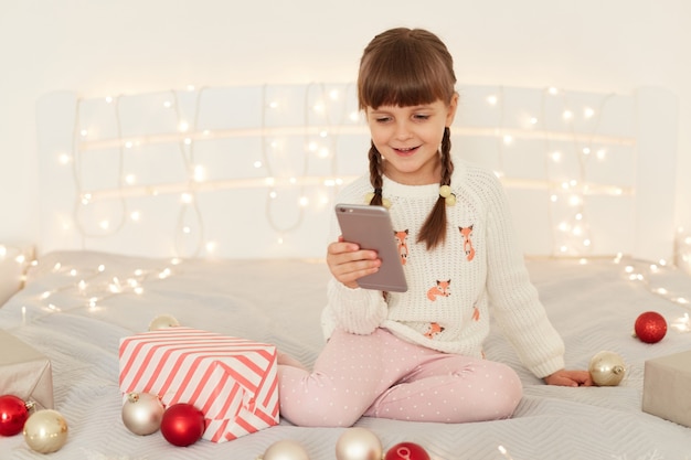 Photo d'intérieur d'une joyeuse enfant aux cheveux noirs avec des nattes tenant un téléphone portable dans les mains et prenant un selfie, regardant la caméra du téléphone intelligent et souriant joyeusement, félicitant Noël.