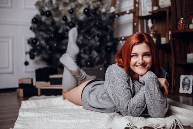 Photo d'intérieur de mode de la belle jeune femme aux cheveux rouges et au sourire charmant, porte un cardigan tricoté confortable, posant à côté de l'arbre de Noël et présente un gâteau au chocolat avec des astérisques