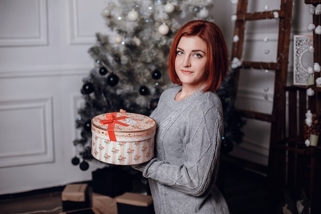 Photo d'intérieur de mode de la belle jeune femme aux cheveux rouges et au sourire charmant, porte un cardigan tricoté confortable, posant à côté de l'arbre de Noël et présente un gâteau au chocolat avec des astérisques