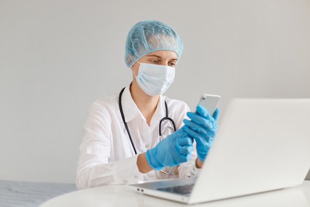 Photo intérieure d'une femme médecin thérapeute concentrée portant une robe, un masque chirurgical, une casquette médicale et des gants, travaillant devant un ordinateur, tenant un téléphone intelligent dans les mains, composant le numéro.