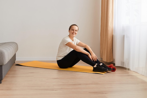 Photo intérieure d'une femme souriante et heureuse faisant de l'exercice à la maison, prenant une pause, se reposant et regardant la caméra avec une expression positive, assise sur un tapis de yoga dans le salon, portant des vêtements de sport.