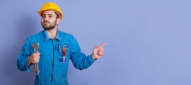 Photo intérieure horizontale de jeune constructeur confiant et beau avec barbe, montrant la direction avec l'index