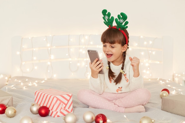 Photo intérieure d'une petite fille adorable et excitée portant un pull décontracté blanc assis sur le lit avec un téléphone portable dans les mains, les poings serrés, célébrant la victoire, regardant l'écran du gadget, ayant un appel vidéo.