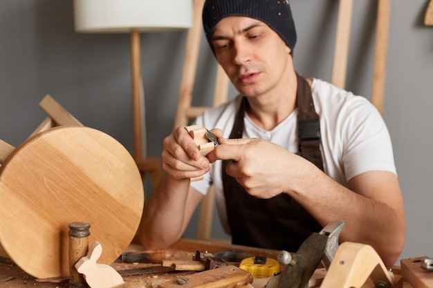 Photo d'un jeune adulte charpentier bricoleur portant un t-shirt blanc, une casquette noire et un tablier marron coupant un morceau de bois incurvé par un coupeur de bois fabriquant des jouets en bois