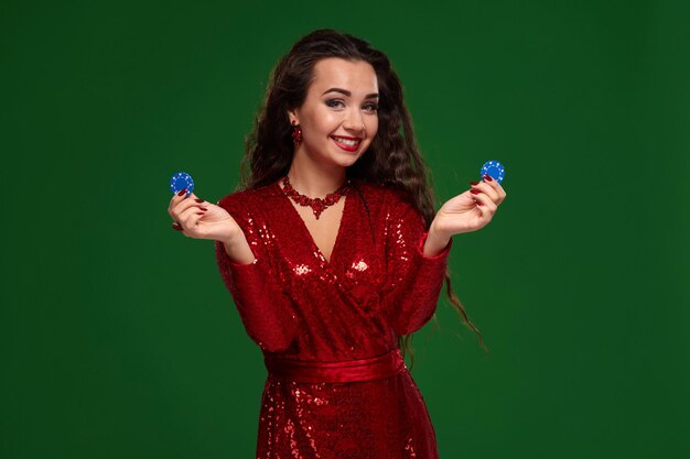 Photo De Jeune Belle Femme Brune Chanceuse Sexy Dans Une Robe De Soirée Rouge Brillante Tient Quelques Jetons De Poker Dans Ses Mains. Fond Vert, Chromakey, Industrie Du Jeu