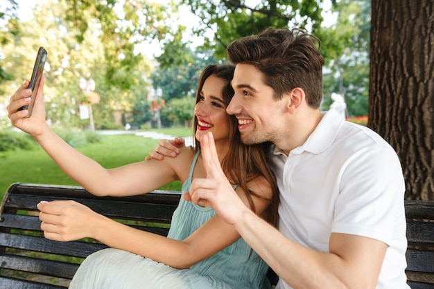 Photo de jeune couple aimant mignon assis à l'extérieur tout en faisant selfie par téléphone mobile montrant le geste de paix