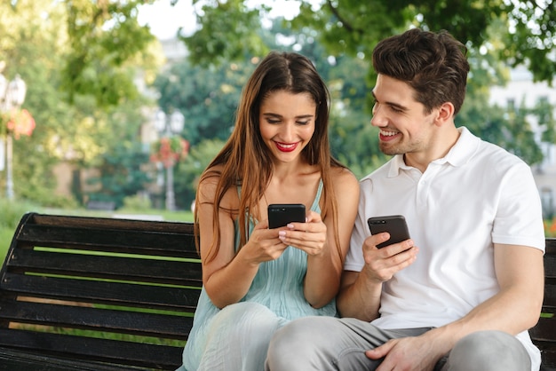 Photo de jeune couple aimant mignon assis à l'extérieur tout en utilisant des téléphones mobiles