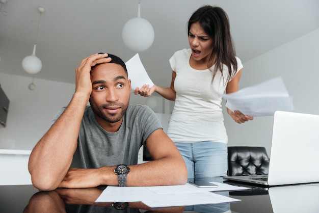 Photo d'un jeune couple d'amoureux discutant des factures domestiques à la maison. Femme criant à l'homme tout en tenant des documents.