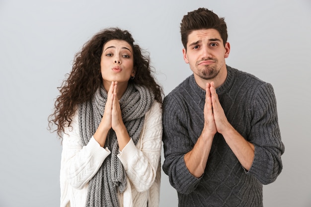 Photo de jeune couple homme et femme priant avec les paumes ensemble, isolé sur mur gris